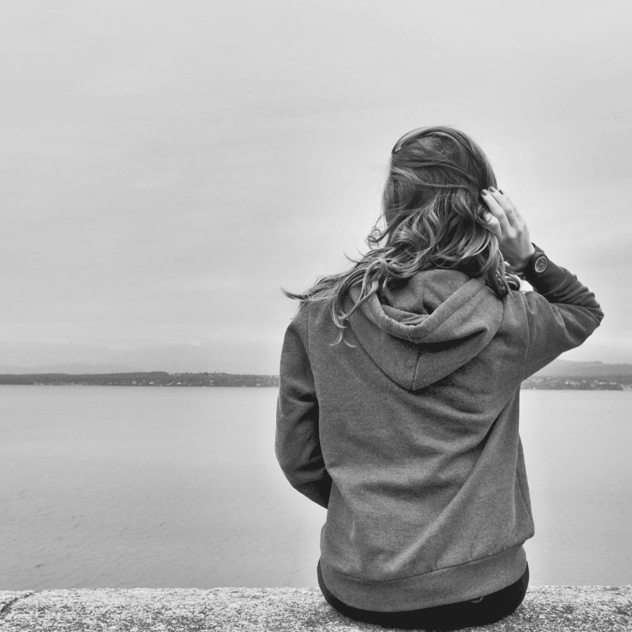 Junge Frau sitzt mit dem Rücken zu uns auf einer Mauer und blickt auf einem See