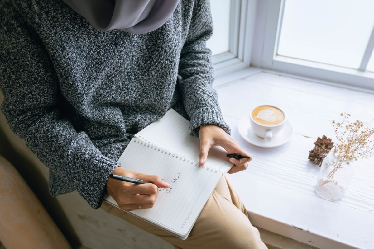Frau schreibt in ein Notizbuch bei einer Tasse Kaffee am Fenster