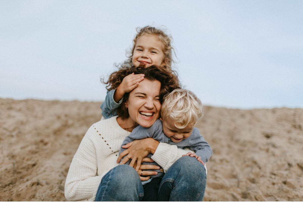 Unser Umfeld prägt unseren Blick auf die Welt Mutter sitzt mit ihren beiden Kindern lachend am Strand.